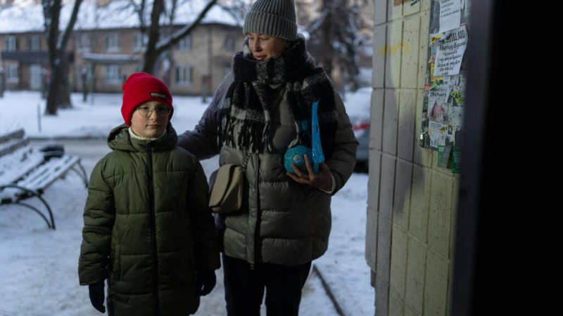 A woman and child in winter attire stand outside in a snowy urban area in Ukraine.